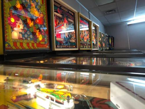 A row of colorful pinball machines with illuminated backglass and shiny playfields in a well-lit arcade.