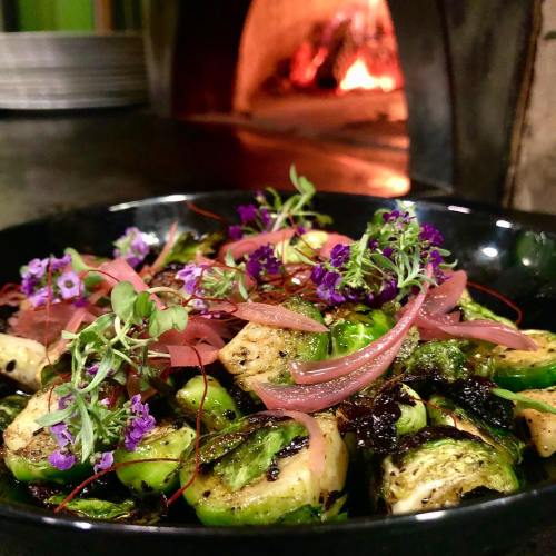 A black bowl filled with grilled Brussels sprouts, pickled onions, and edible flowers, with a warm fireplace in the background.