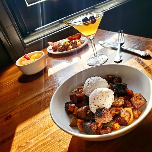 A bowl of poached eggs on roasted potatoes, with a cocktail and fruit in the background on a wooden table.