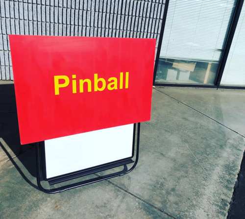 A red sign with yellow text that reads "Pinball," placed on a concrete surface outside a building.