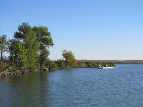 A serene lake scene with trees on the shore and a small white dock extending into the water under a clear blue sky.