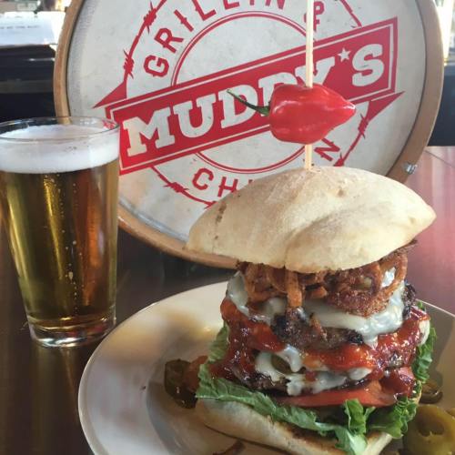 A towering burger with layers of meat, toppings, and a chili pepper, served with a glass of beer in a casual setting.