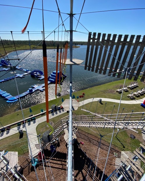 View from the top of a climbing structure overlooking a lake with floating obstacles and a grassy area.