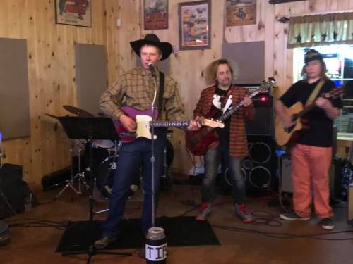 A band performs on stage in a rustic venue, featuring a guitarist, bassist, and a singer with a cowboy hat.