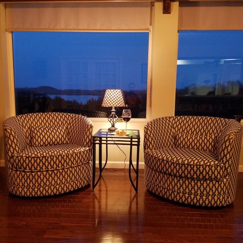 Two patterned chairs beside a small table with a lamp and a glass of wine, set against a window at dusk.