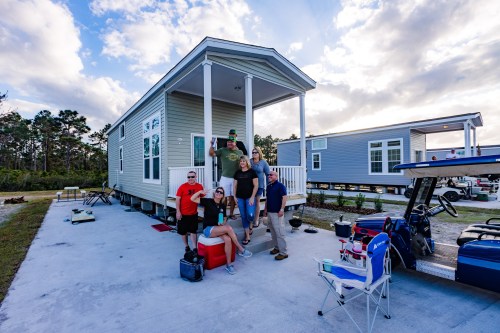 A group of six people poses in front of a modern mobile home, with a golf cart nearby and a scenic sky in the background.