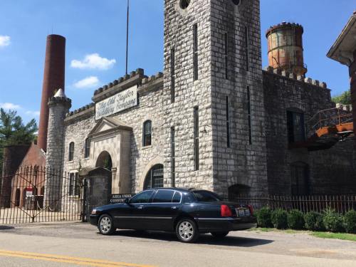 A black car parked in front of a stone building with a tower and industrial features under a clear blue sky.