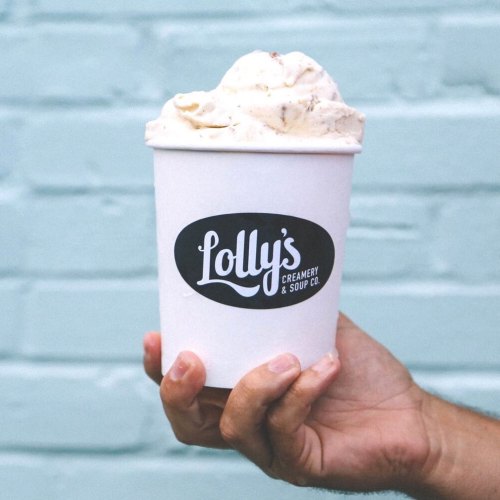 A hand holding a cup of ice cream from Lolly's Creamery & Soup Co. against a light blue brick wall.