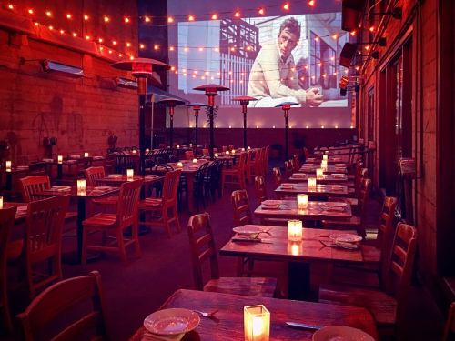 A cozy outdoor dining area with wooden tables, candles, and a movie screen showing a man, illuminated by string lights.
