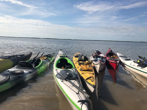 A group of colorful kayaks lined up on a calm water surface under a partly cloudy sky.