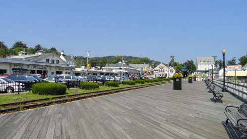 A sunny boardwalk lined with benches, shops, and parked cars, with green bushes and hills in the background.