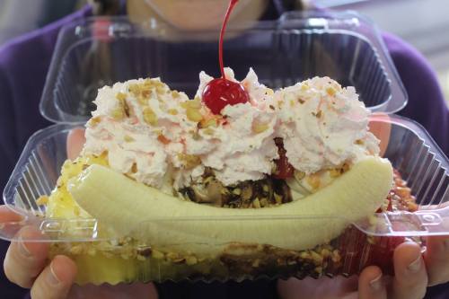 A person holding a plastic container with a banana split topped with whipped cream, nuts, and a cherry.