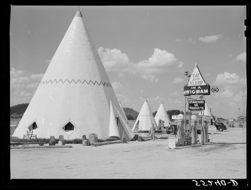Historic image of white teepees and a sign for "Eat and Sleep in a Wigwam" with vintage gas pumps nearby.