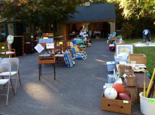 A yard sale with various items for sale, including furniture, boxes, and sports equipment, set in a driveway.