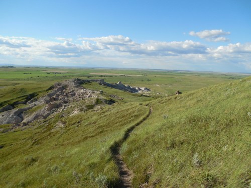 A winding path leads through lush green hills under a blue sky with scattered clouds, showcasing a vast landscape.