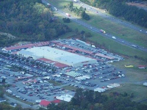 Aerial view of a large warehouse surrounded by a parking lot and nearby highway, with various vehicles and a circus tent.