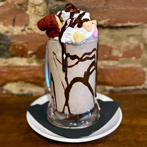 A tall glass of milkshake topped with whipped cream, chocolate drizzle, and a piece of red velvet cake.