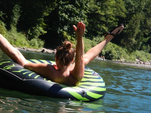 A person relaxes on a green and black inflatable tube in a river, with arms raised and legs extended.