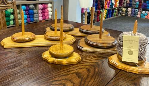 Wooden yarn holders on a table, with a yarn ball and colorful yarns displayed in the background.