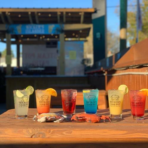 A row of colorful cocktails on a wooden table, with a crab and a seafood restaurant backdrop.