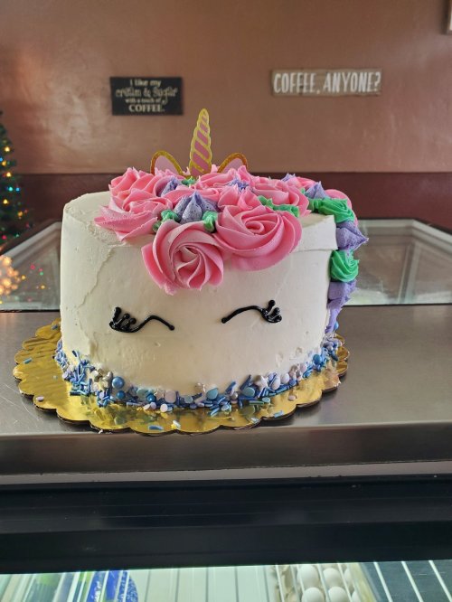 A whimsical unicorn cake with pink roses, a golden horn, and colorful decorations, displayed in a bakery.