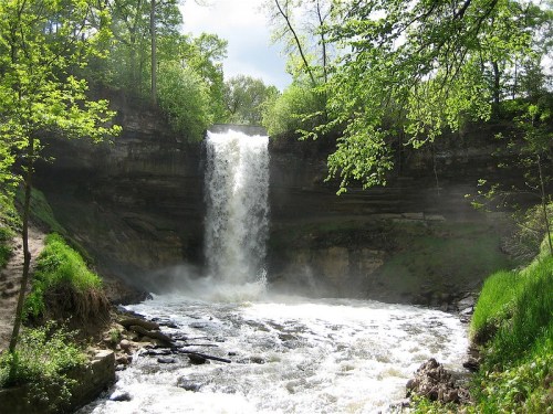 A cascading waterfall surrounded by lush green trees and rocky terrain, with mist rising from the water below.