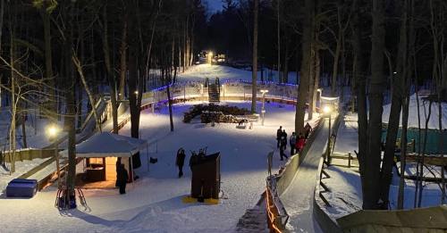 A snowy forest scene at night with illuminated pathways, people walking, and a cozy gathering area.
