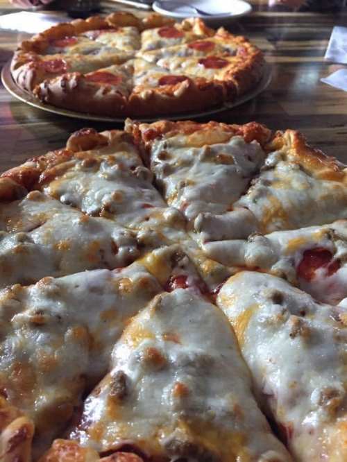 Close-up of a cheesy pizza with pepperoni and sausage, with another pizza in the background on a wooden table.