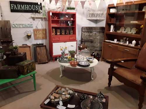 A cozy vintage shop interior featuring rustic furniture, decor items, and a central coffee table with various collectibles.