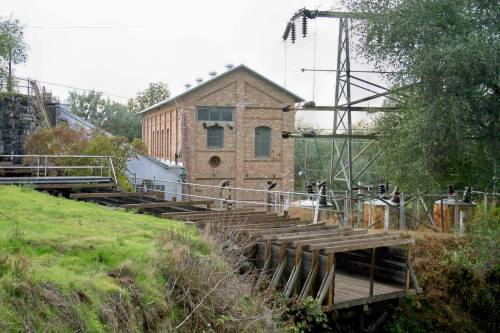Historic brick building near power lines, surrounded by greenery and wooden structures, showcasing industrial architecture.
