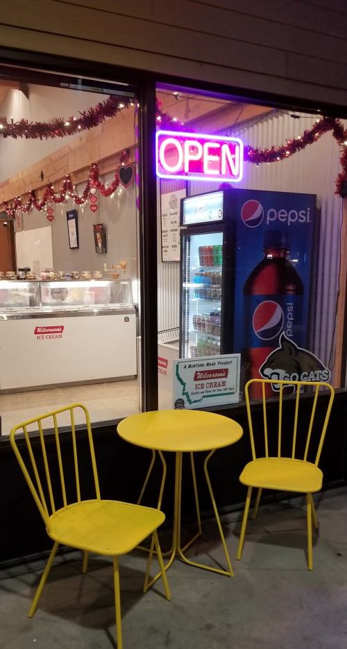 A brightly lit shop window with an "OPEN" sign, featuring a small yellow table and chairs outside.