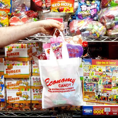 A hand holds a white Economy Candy bag filled with colorful candies, in front of a shelf stocked with various sweets.