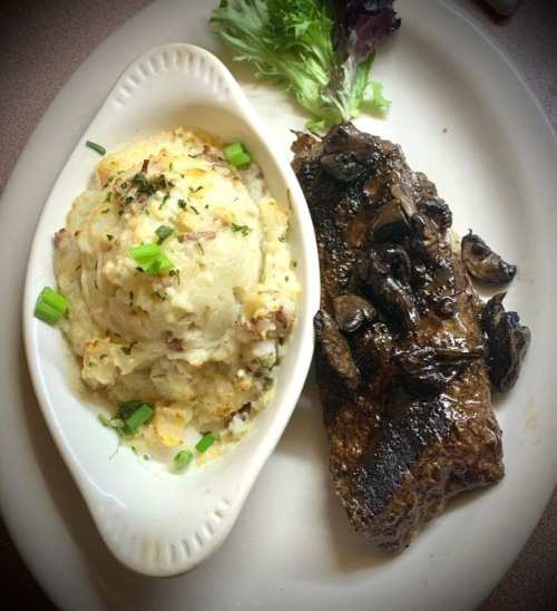 A plate featuring a grilled steak with sautéed mushrooms and a side of creamy mashed potatoes garnished with green onions.