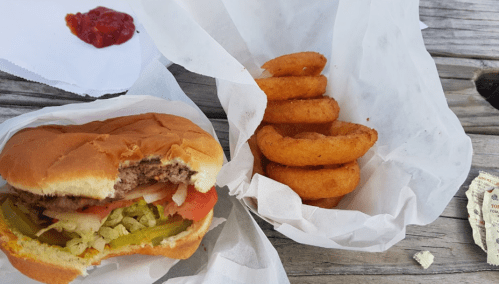 A half-eaten hamburger with lettuce, tomato, and pickles next to a serving of onion rings and a packet of ketchup.