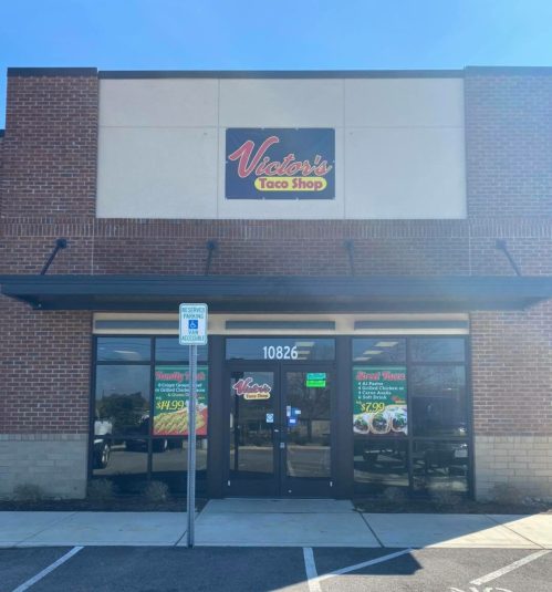 Exterior of a taco shop with a sign reading "Victor's Taco Shop," featuring large windows and a parking area.