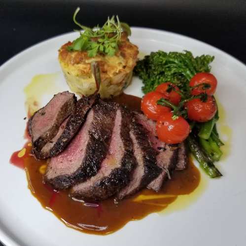 Sliced beef with a rich sauce, served with roasted tomatoes, broccolini, and a savory potato cake.