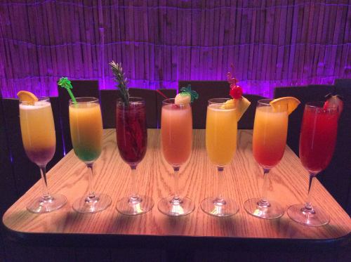 A colorful array of seven cocktails in tall glasses, each garnished with fruit and decorative elements, on a wooden table.