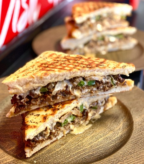 A close-up of a grilled sandwich with layers of meat and vegetables, served on a wooden plate.