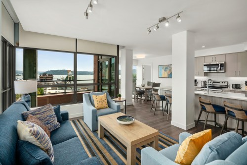Bright, modern living room with blue sofas, a coffee table, and a view of the water through large windows.