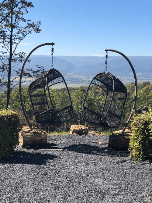 Two hanging wicker chairs on a stand, overlooking a scenic mountain view under a clear blue sky.