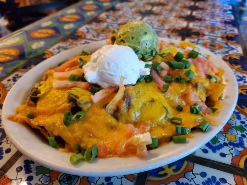 A plate of nachos topped with melted cheese, guacamole, green onions, and diced tomatoes on a colorful tiled table.