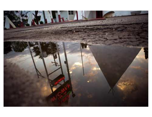 A puddle reflects a retro motel sign and clouds, with a triangular building in the background.
