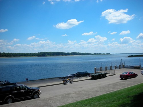 A scenic view of a river under a blue sky with fluffy clouds, featuring parked cars and a distant shoreline.