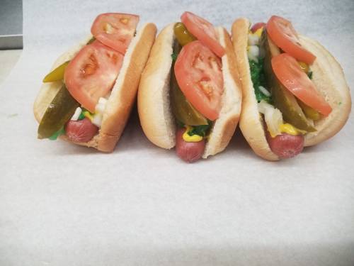 Three hot dogs in buns, topped with lettuce, pickles, tomatoes, and mustard, arranged on a white surface.