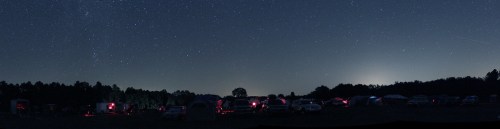 A panoramic view of a campsite at night, with tents and cars illuminated by red lights under a starry sky.