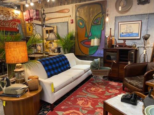 A cozy vintage shop interior featuring a white sofa, colorful artwork, various decor items, and a patterned rug.