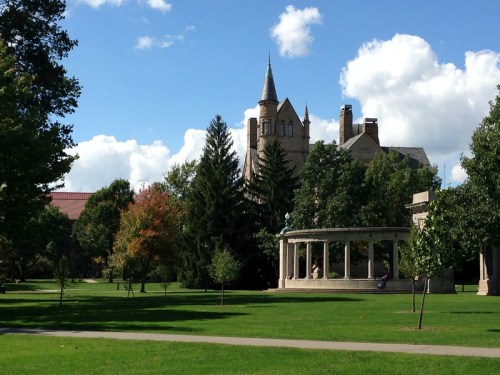 A scenic view of a park featuring a gazebo, trees, and a historic building with a tower under a blue sky.