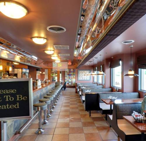 A modern diner interior with booths, a long counter, and colorful decor, featuring a "Please Wait To Be Seated" sign.