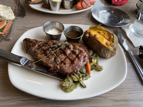 A grilled steak with herbs, served with a baked potato topped with cheese and vegetables on the side.