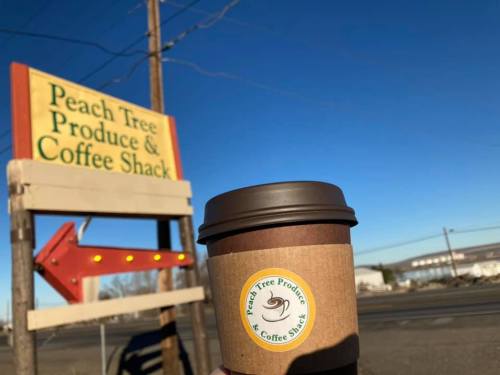 A hand holds a coffee cup in front of the Peach Tree Produce & Coffee Shack sign against a clear blue sky.